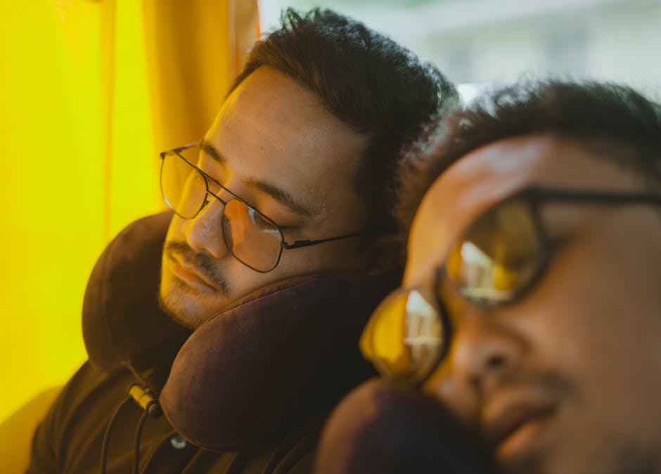 Two men wearing eyeglasses sleep on a bus, using neck pillows for comfort during the journey.