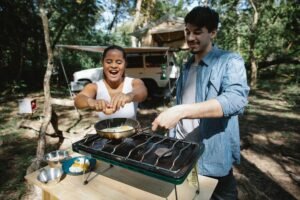Pareja cocinando huevos con un hornillo de camping de gas junto a la tienda de campaña en el bosque