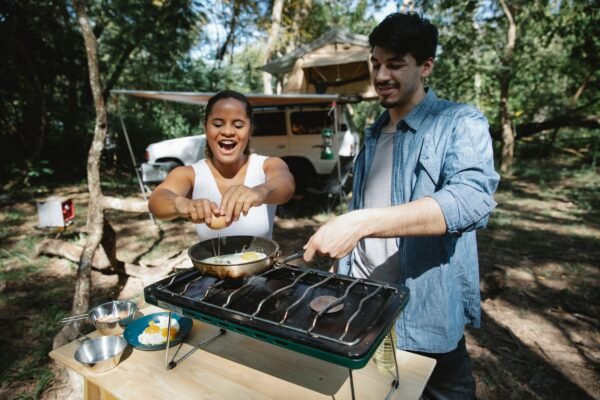 Pareja cocinando huevos con un hornillo de camping de gas junto a la tienda de campaña en el bosque