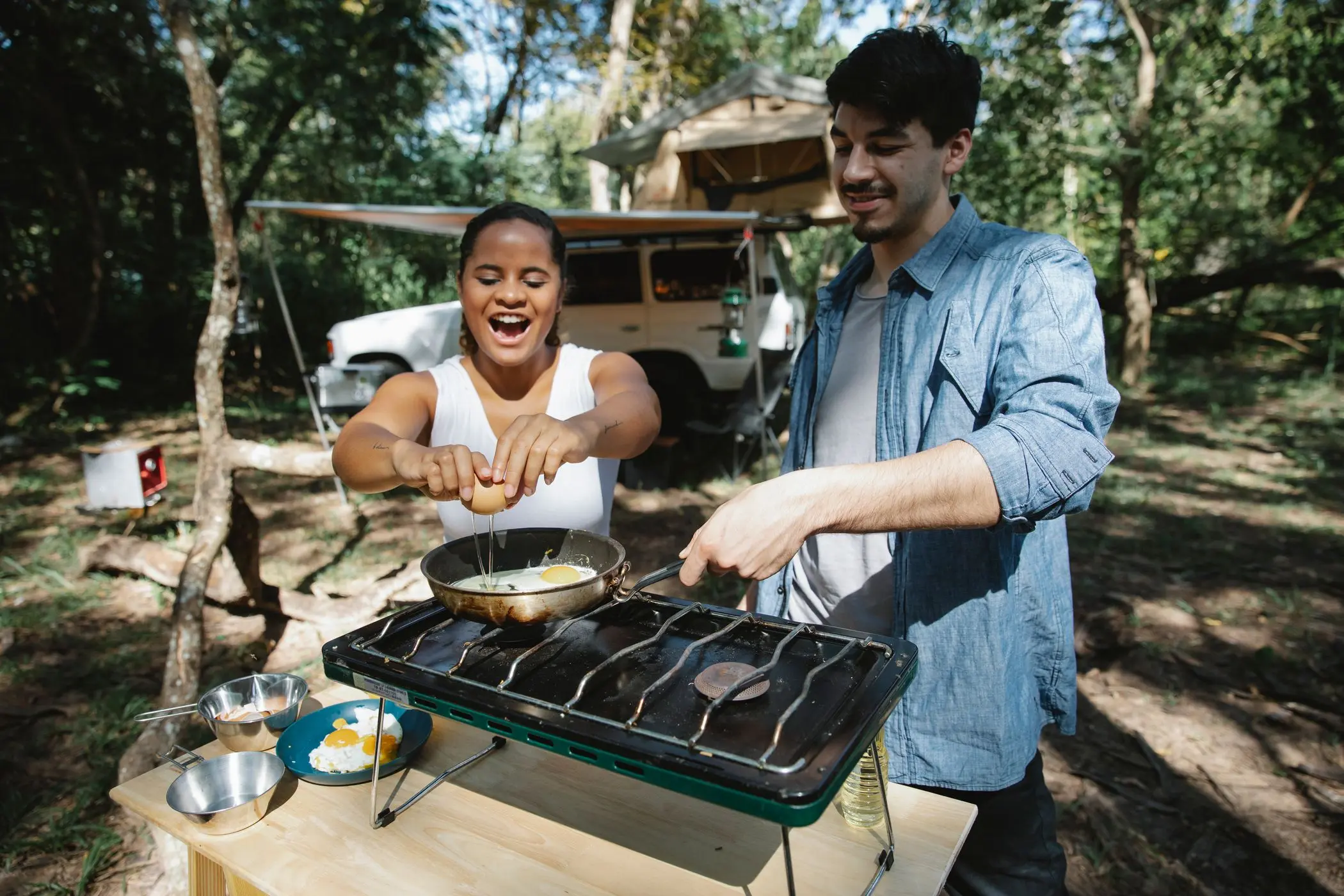 Pareja cocinando huevos con un hornillo de camping de gas junto a la tienda de campaña en el bosque