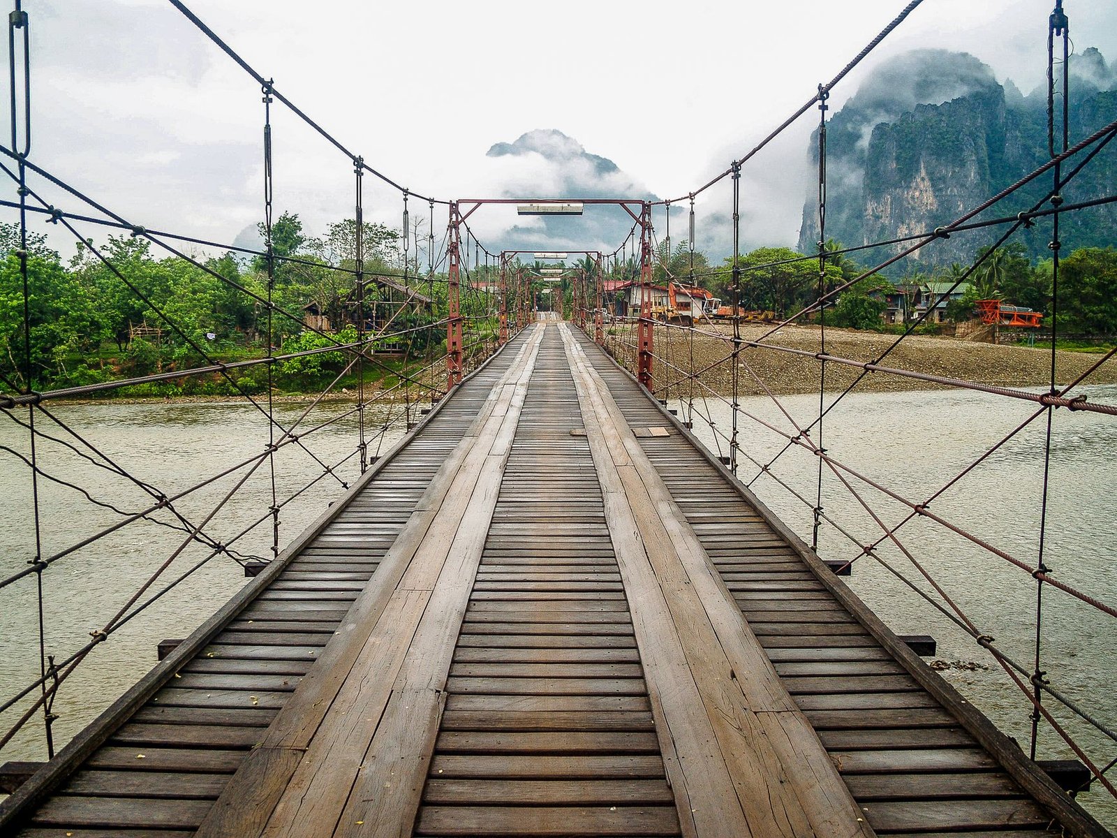 Puente colgante de madera sobre un río con montañas entre la niebla en Vang Vieng